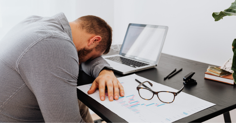 Exhausted office worker with head down on desk beside laptop and data charts, showing signs of burnout and workplace fatigue